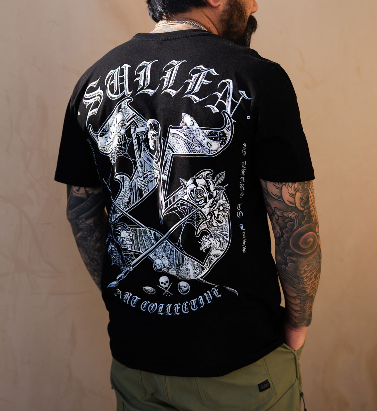 Person wearing a black t-shirt with white graphics and text on the back, standing against a beige wall.