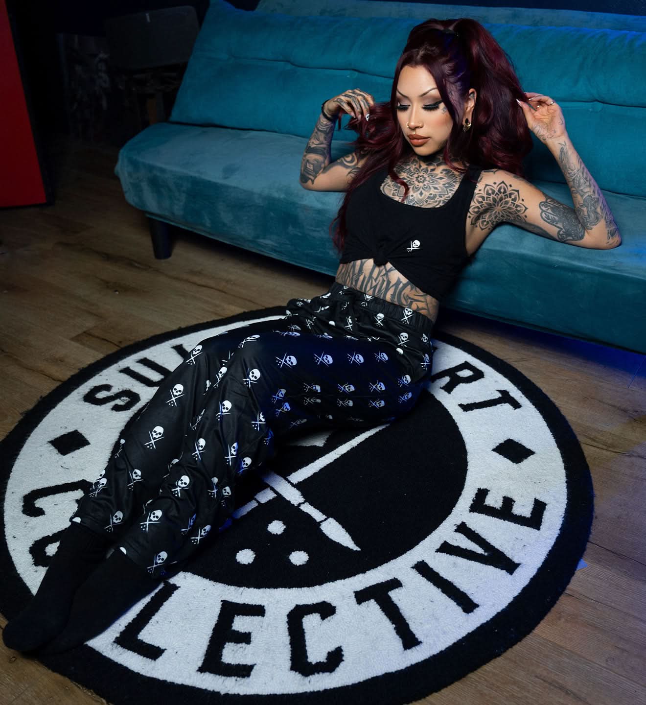 Woman with tattoos sitting on a round rug with text, wearing a black outfit.