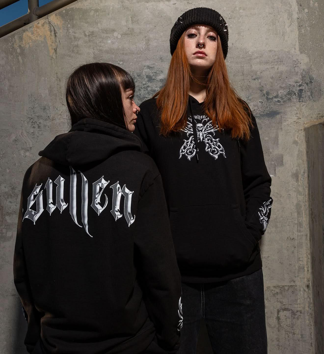 Two people wearing black hoodies with visible branding against a concrete wall.