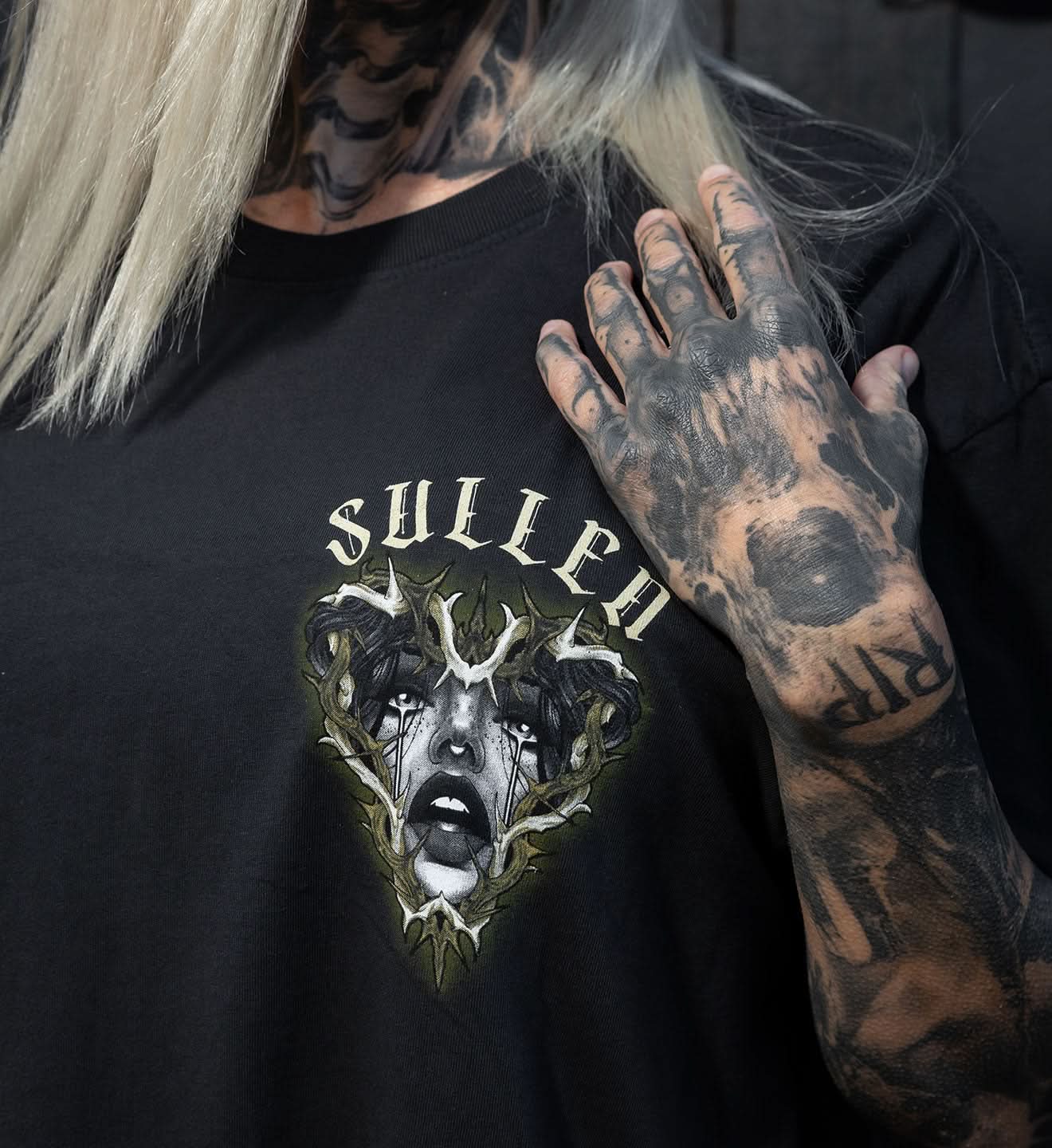 Person wearing a black t-shirt with a graphic design and 'Sullen' branding, holding their tattooed arm.