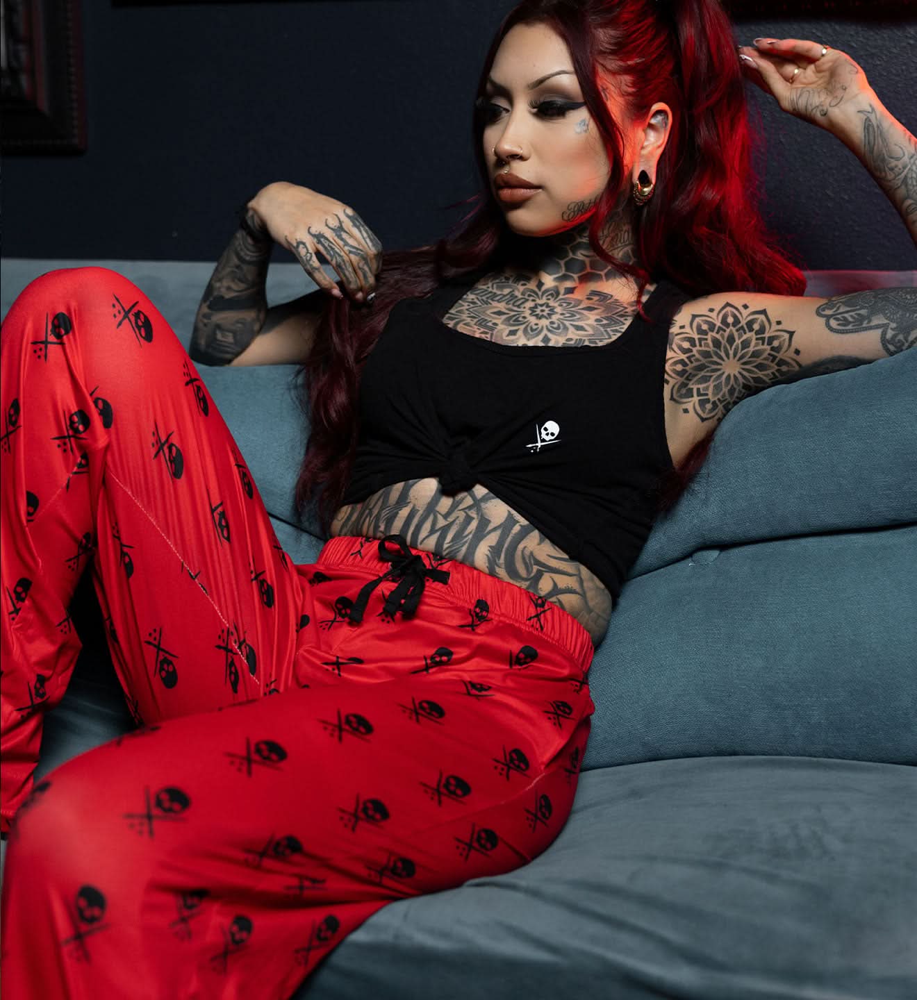 Person with tattoos wearing red pants and a black top sitting on a blue couch.