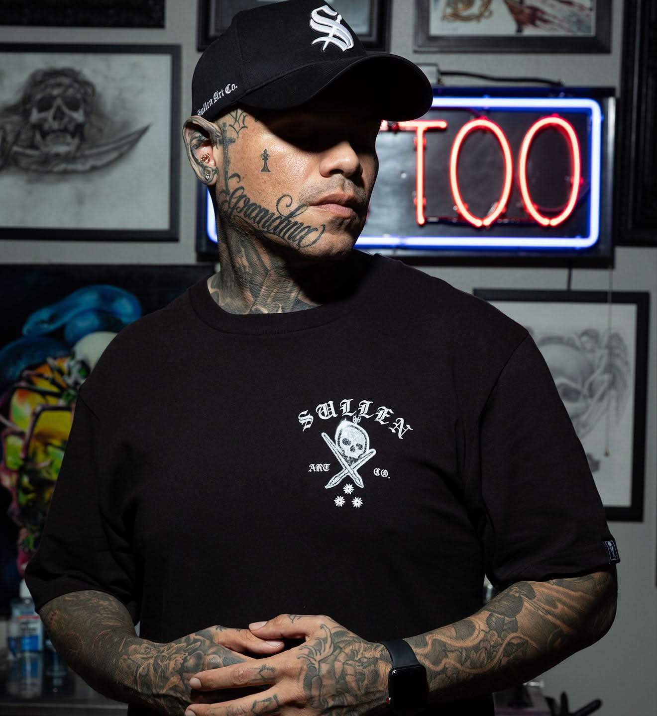 Person wearing a black t-shirt with a logo and text, standing in a tattoo parlor.