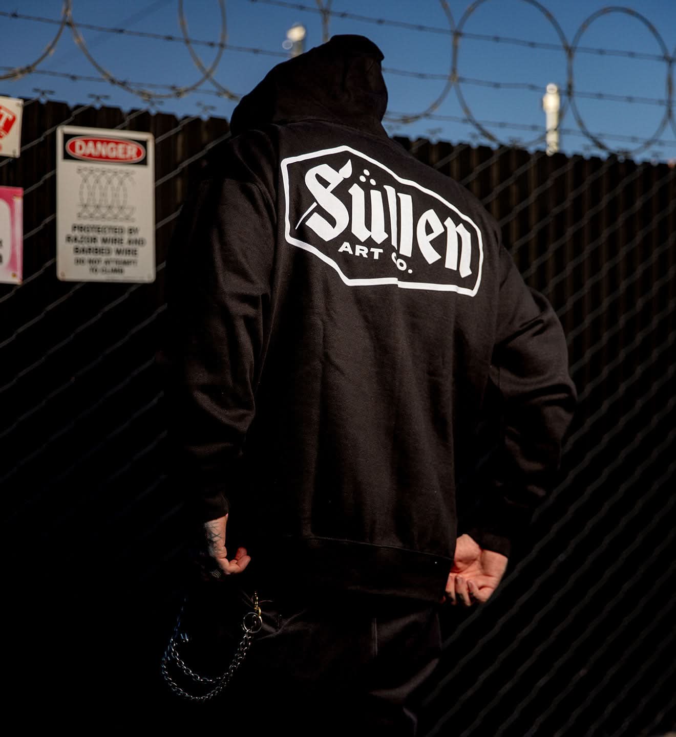 Person wearing a black hoodie with 'Sullen Art Co.' logo against a barbed wire fence.