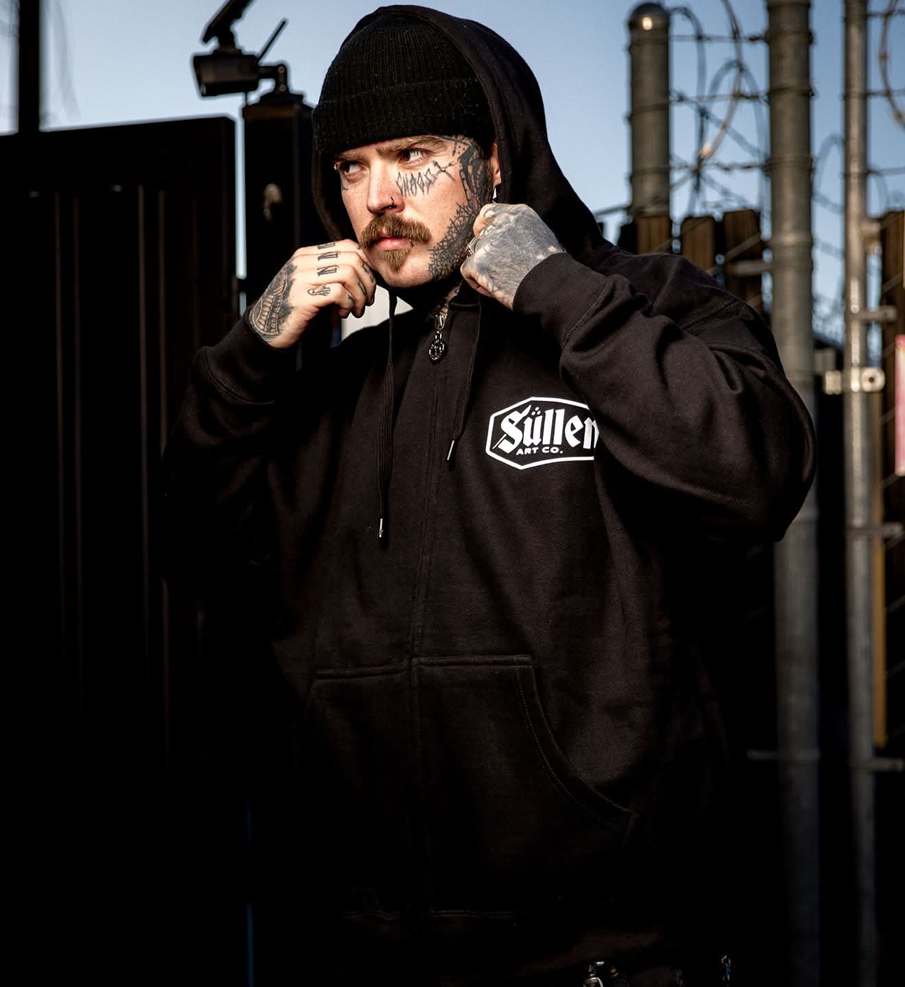 Person wearing a black hoodie with a logo, standing in front of a barbed wire fence.