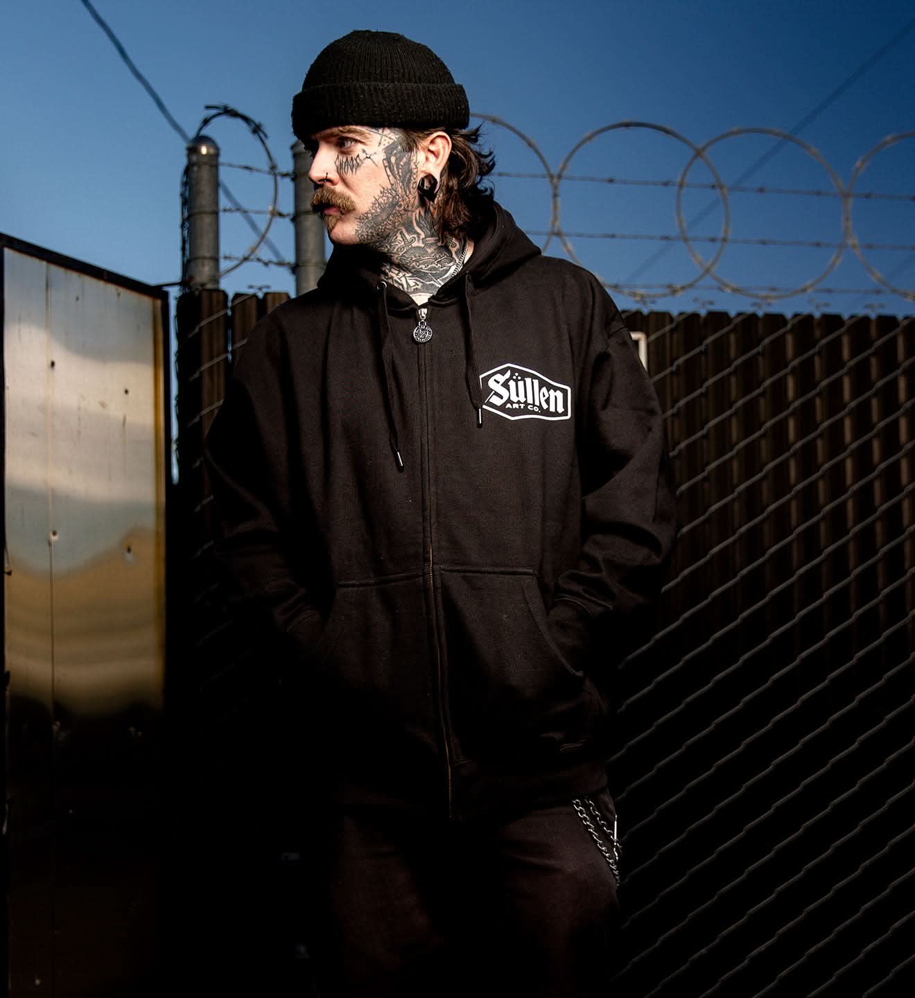 Person wearing a black hoodie with a brand logo, standing in front of a barbed wire fence.