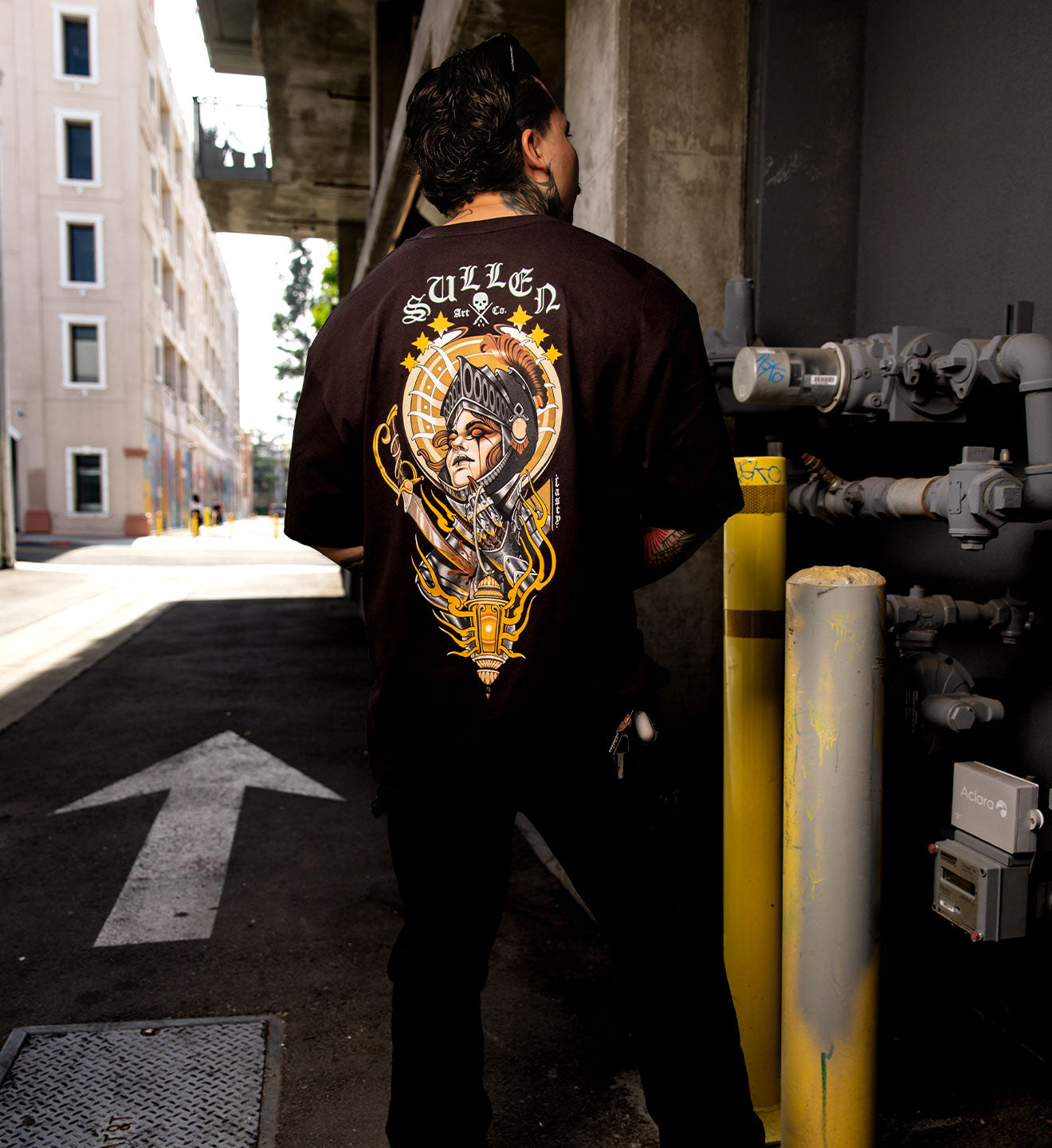 Person wearing a black t-shirt with a graphic design on the back, standing in an urban alleyway.