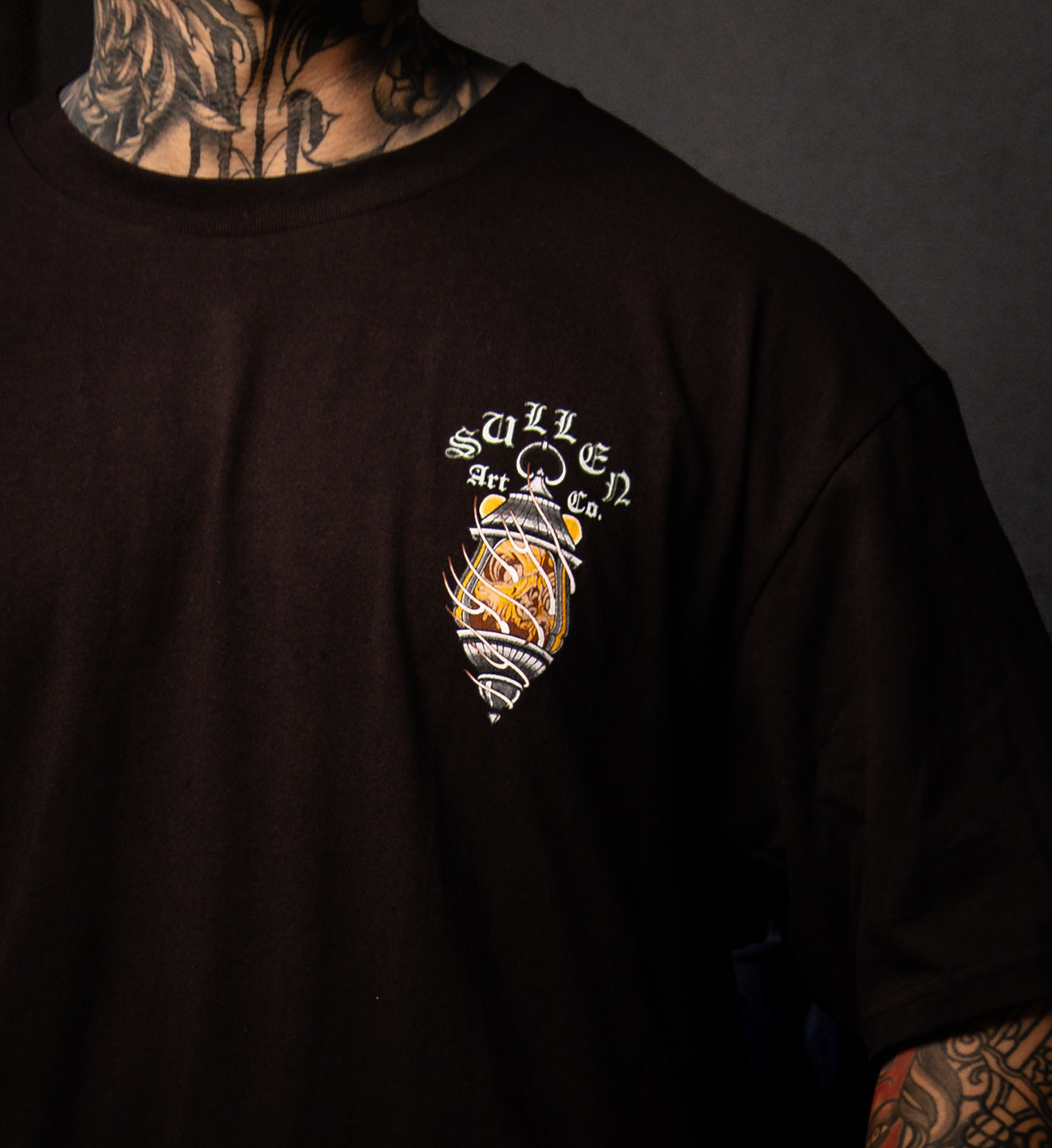 Black t-shirt with a logo on the chest, worn by a person with tattoos.