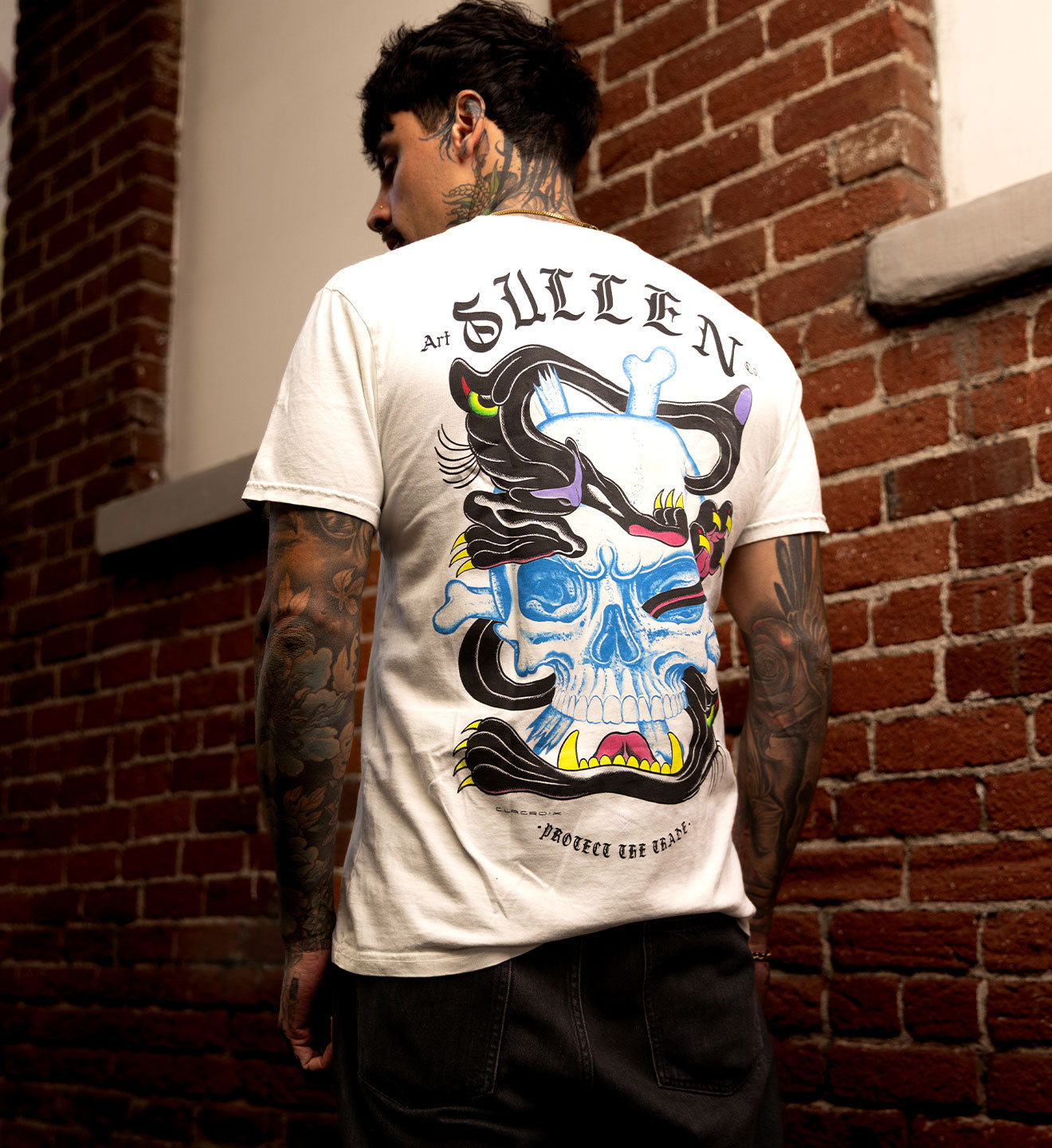 Person wearing a white t-shirt with a colorful skull design and 'Sullen' branding against a brick wall.