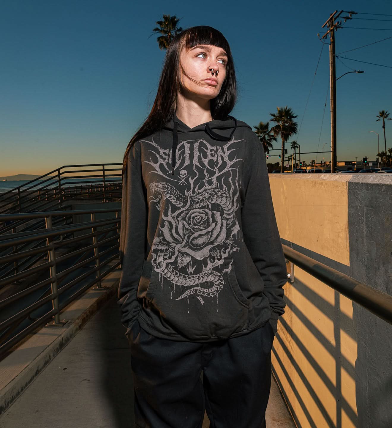 Person wearing a black hoodie with a graphic design on a pier with palm trees in the background