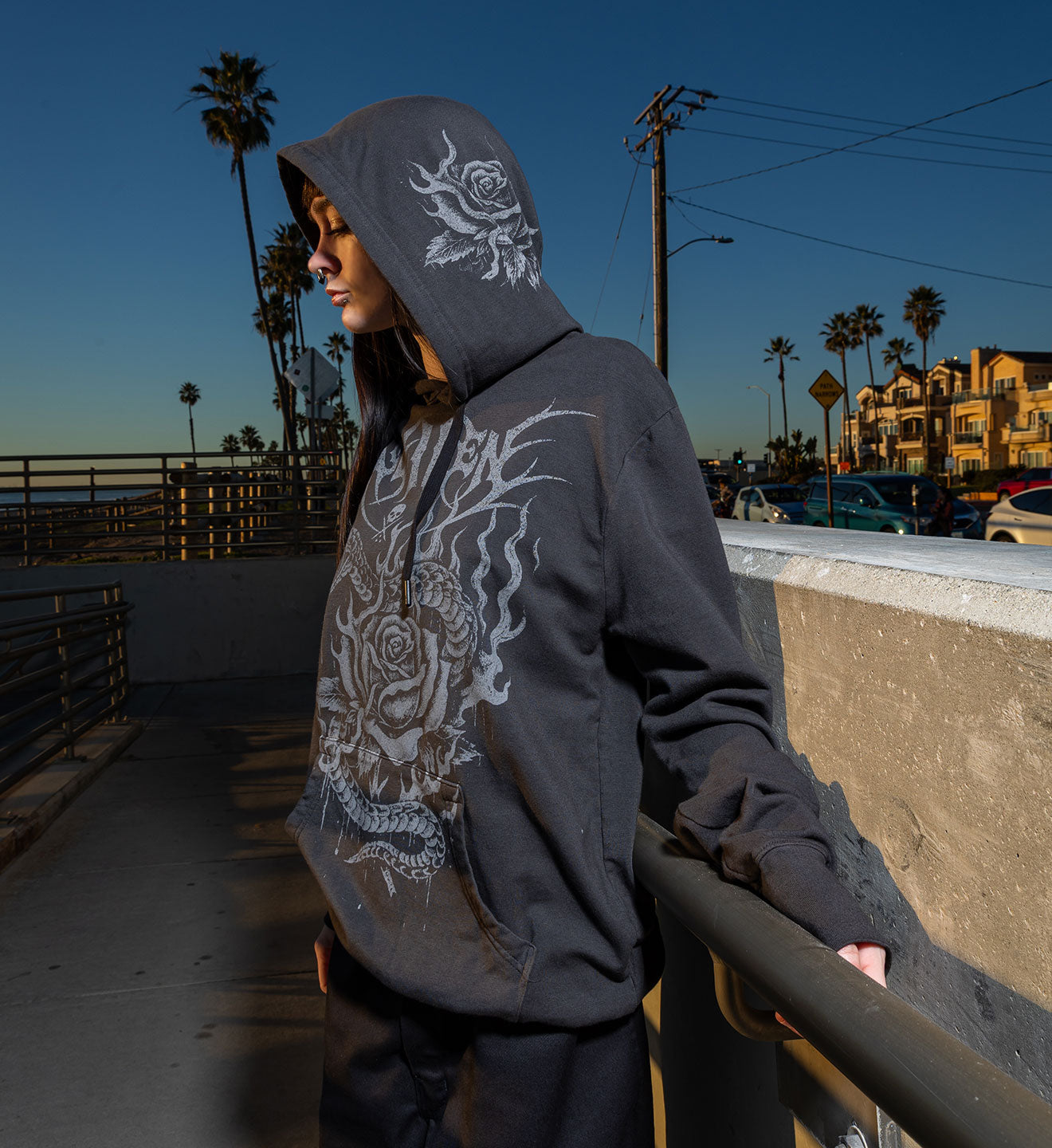 Person wearing a dark hoodie with a white design, standing on a pier with palm trees and buildings in the background.