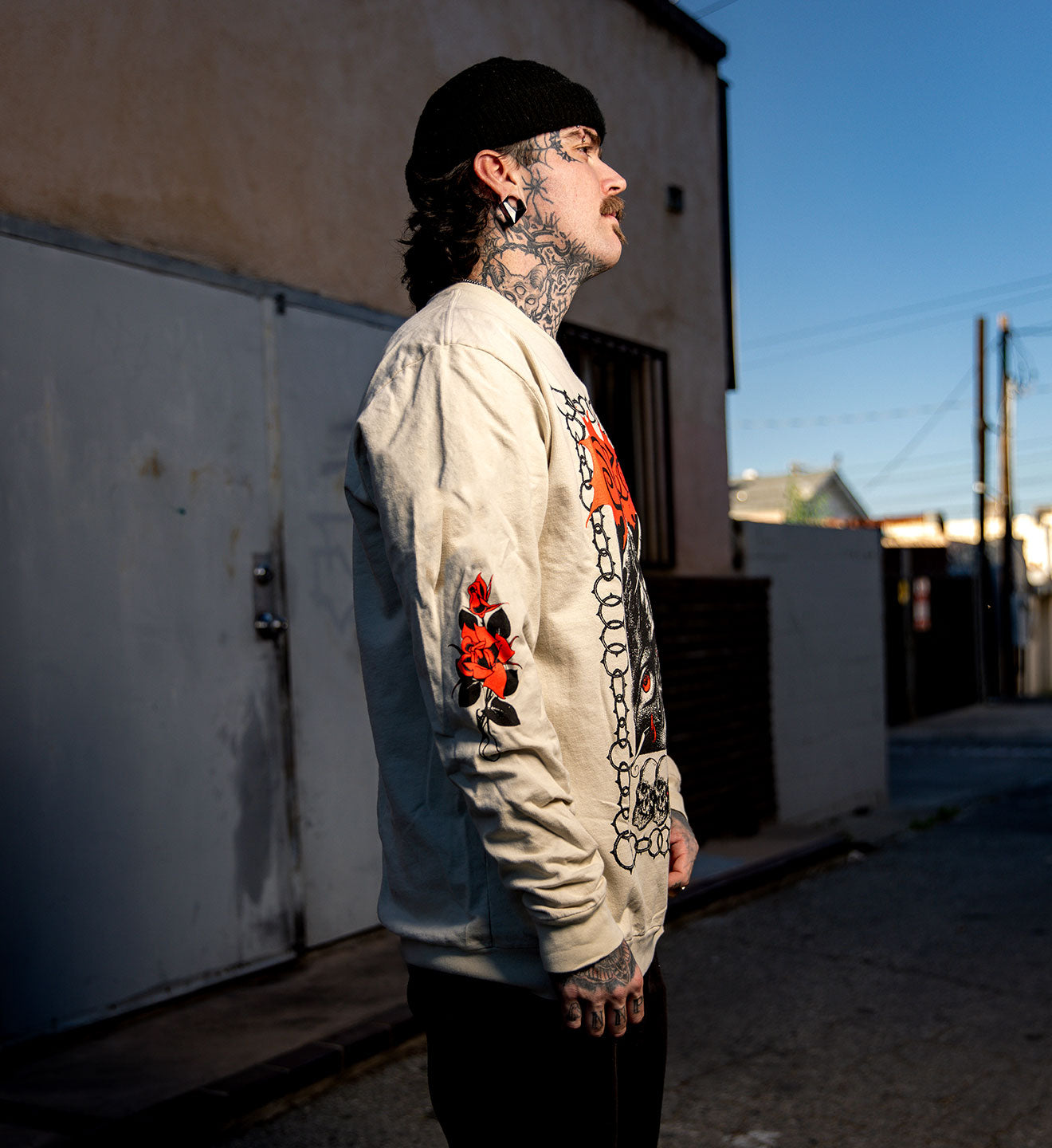 Man with tattoos wearing a light-colored jacket with floral designs standing in an alleyway.