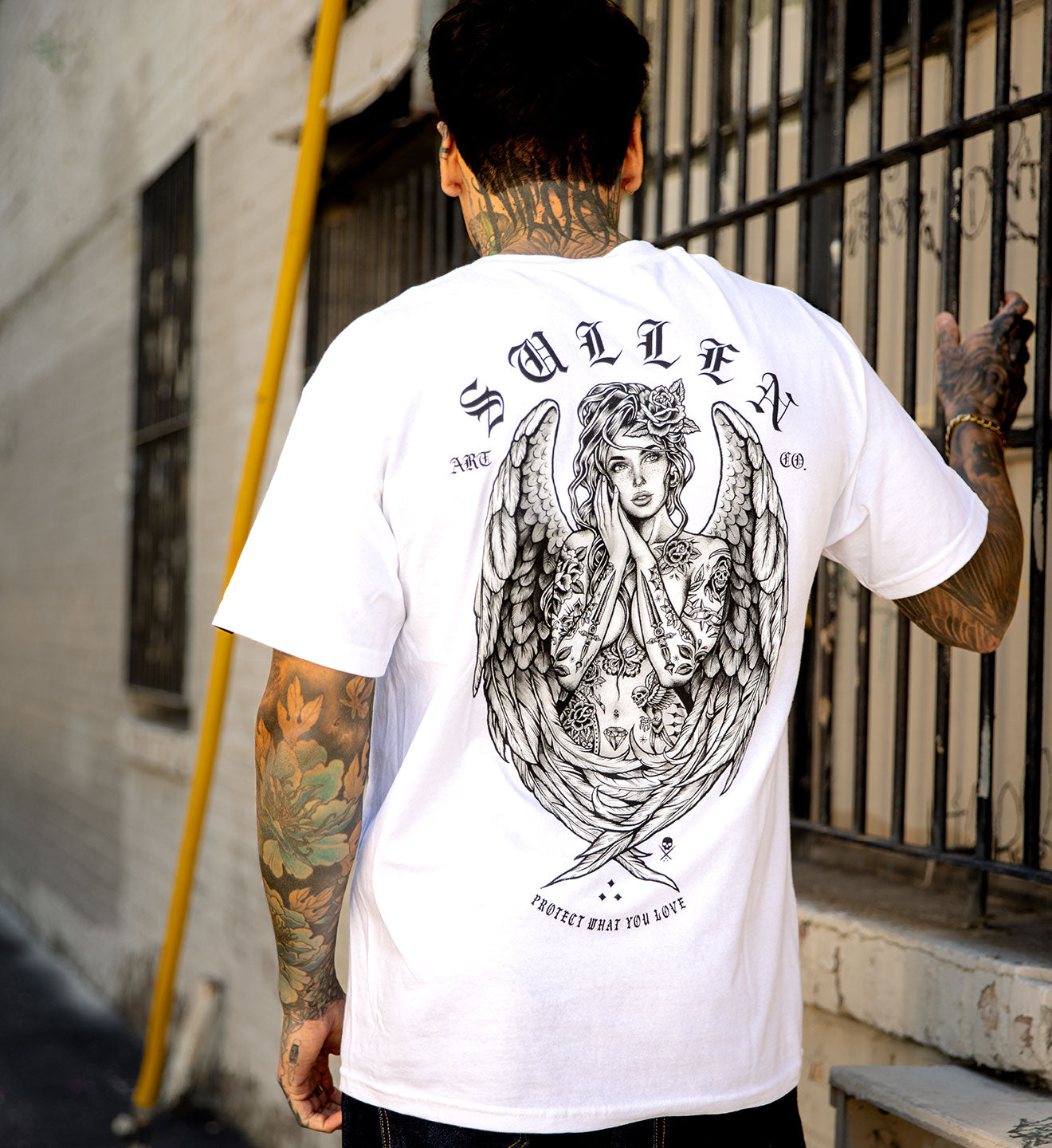 Person wearing a white t-shirt with an angel design on the back, standing against a wall.