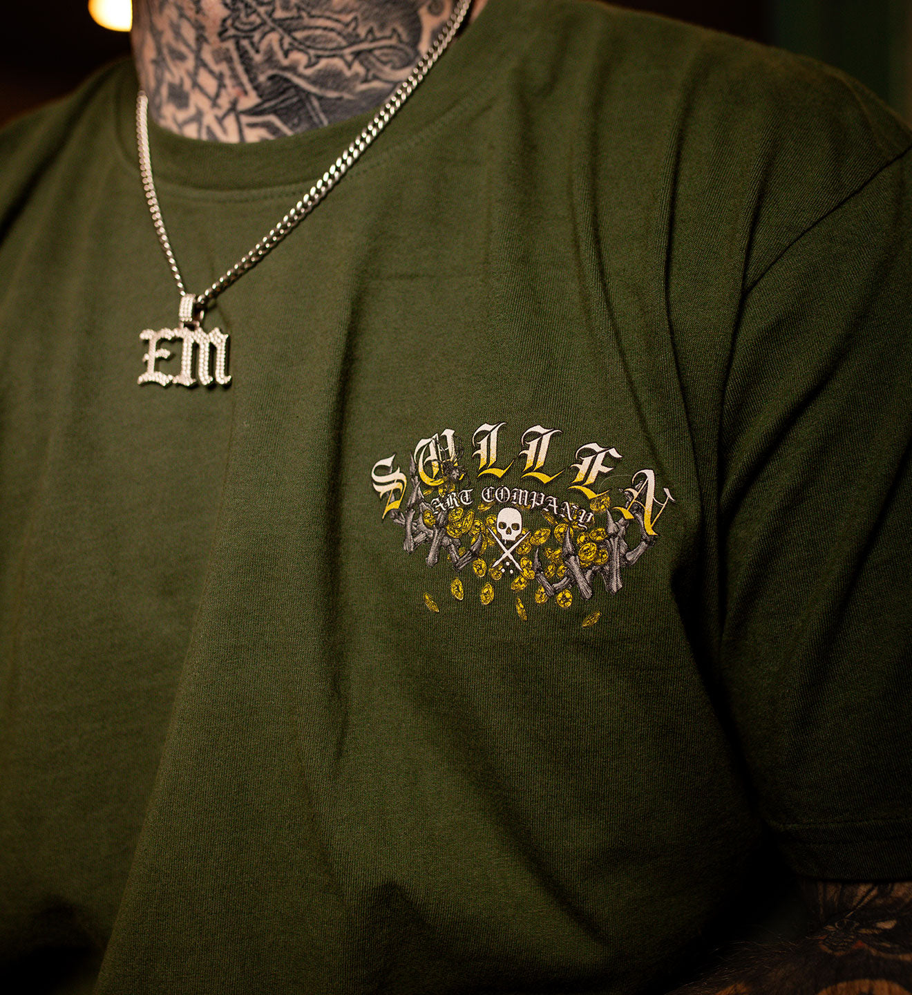 Green t-shirt with 'Sullen art co' logo and tattooed arm wearing a necklace.