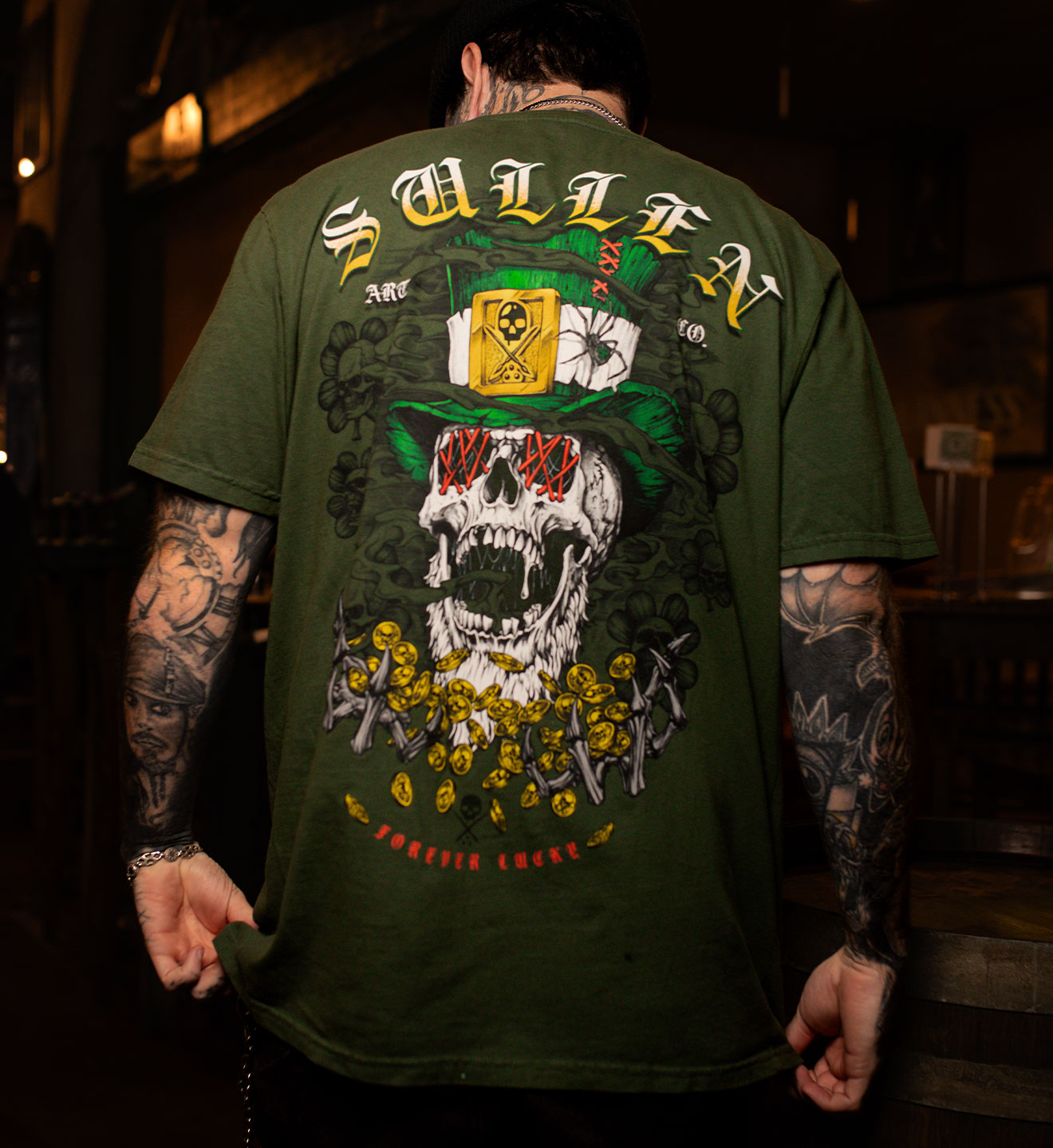 Person wearing a green t-shirt with a skull design and text on the back, standing in a dimly lit room.