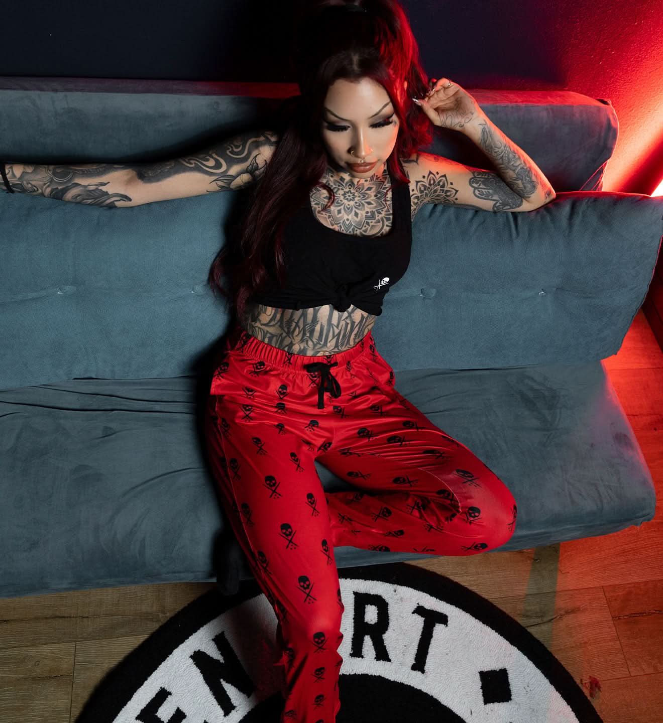 Person with tattoos wearing a black top and red pants sitting on a blue couch.