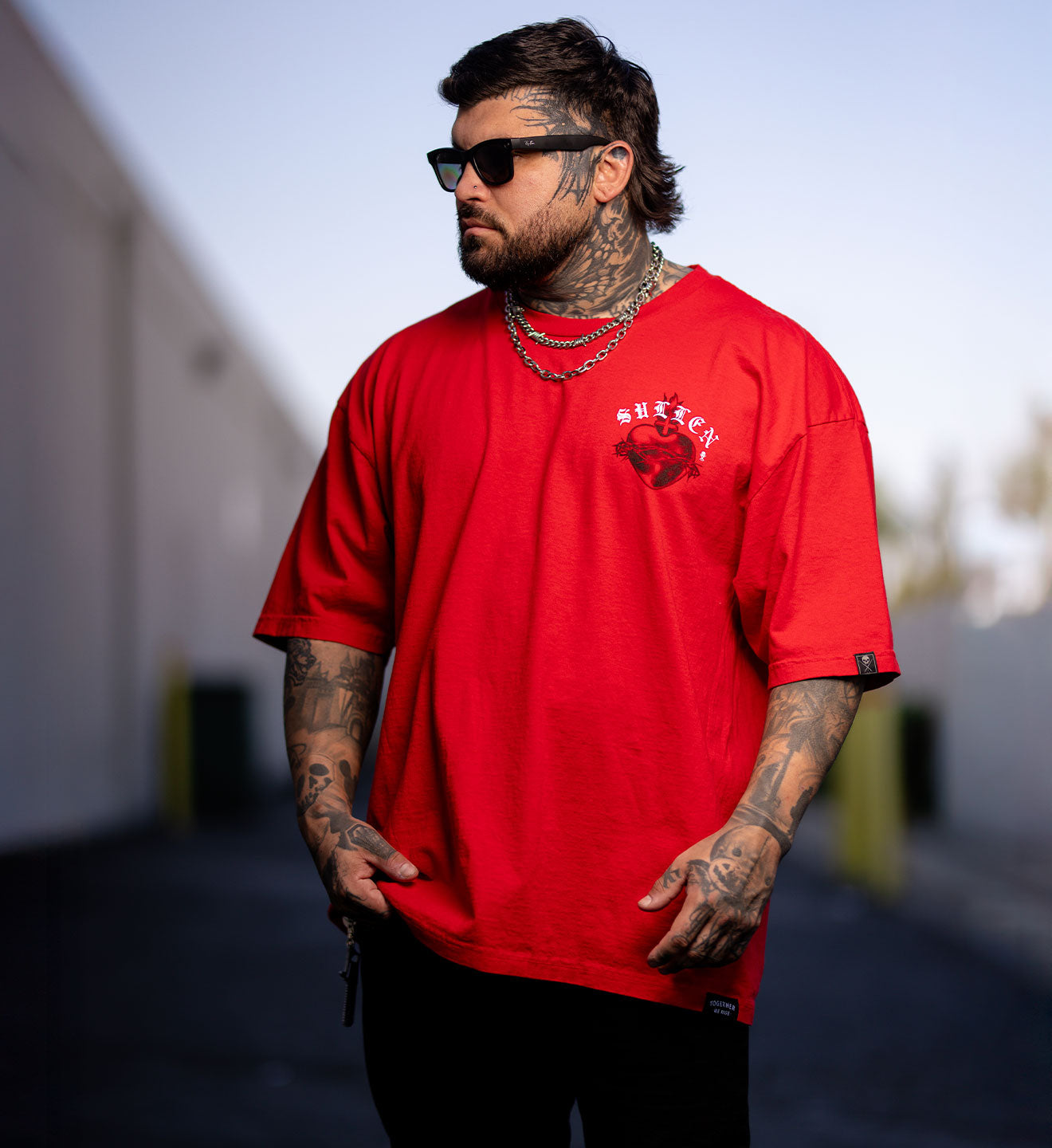 Man wearing a red t-shirt with a logo, sunglasses, and tattoos standing outdoors.