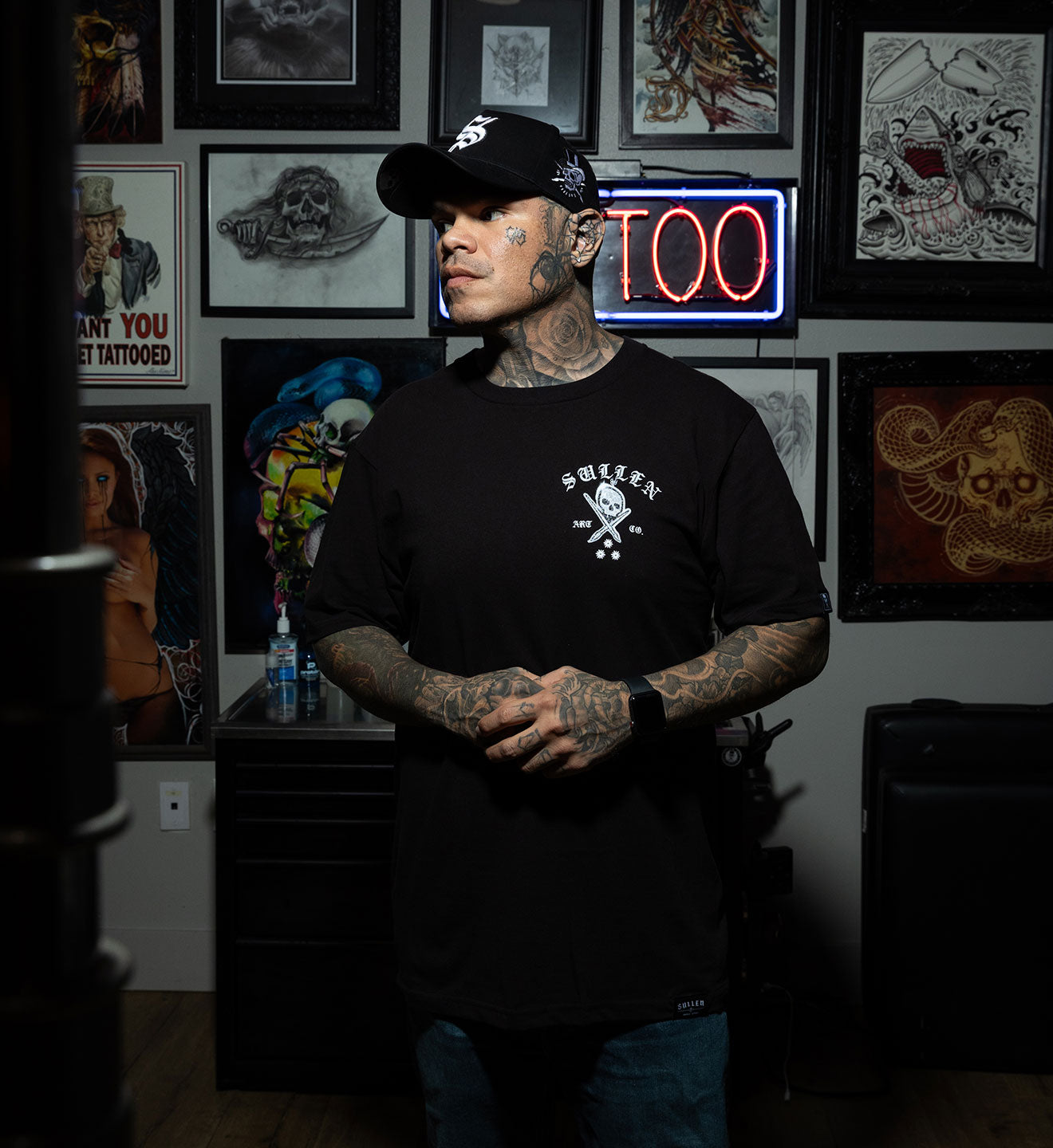 Man with tattoos wearing a black t-shirt and cap in a tattoo parlor with framed artwork on the wall.