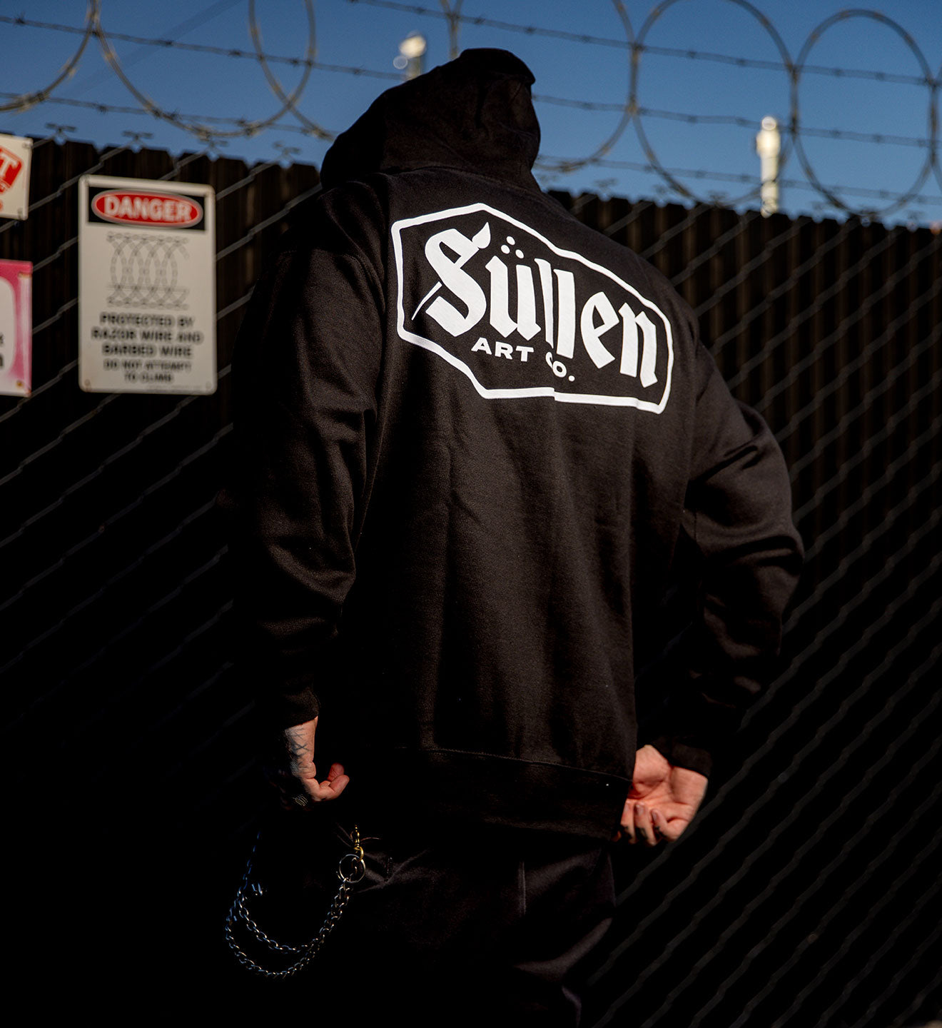Person wearing a black hoodie with 'Sullen Art Co.' logo against a barbed wire fence.