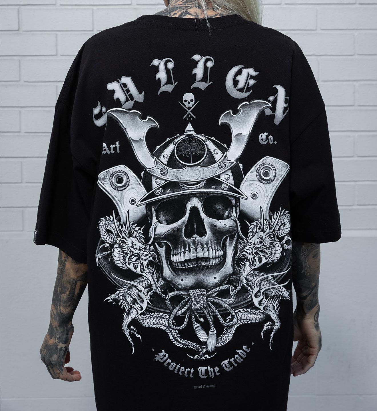 Person wearing a black t-shirt with a detailed skull and crossbones design on the back, against a white brick wall.