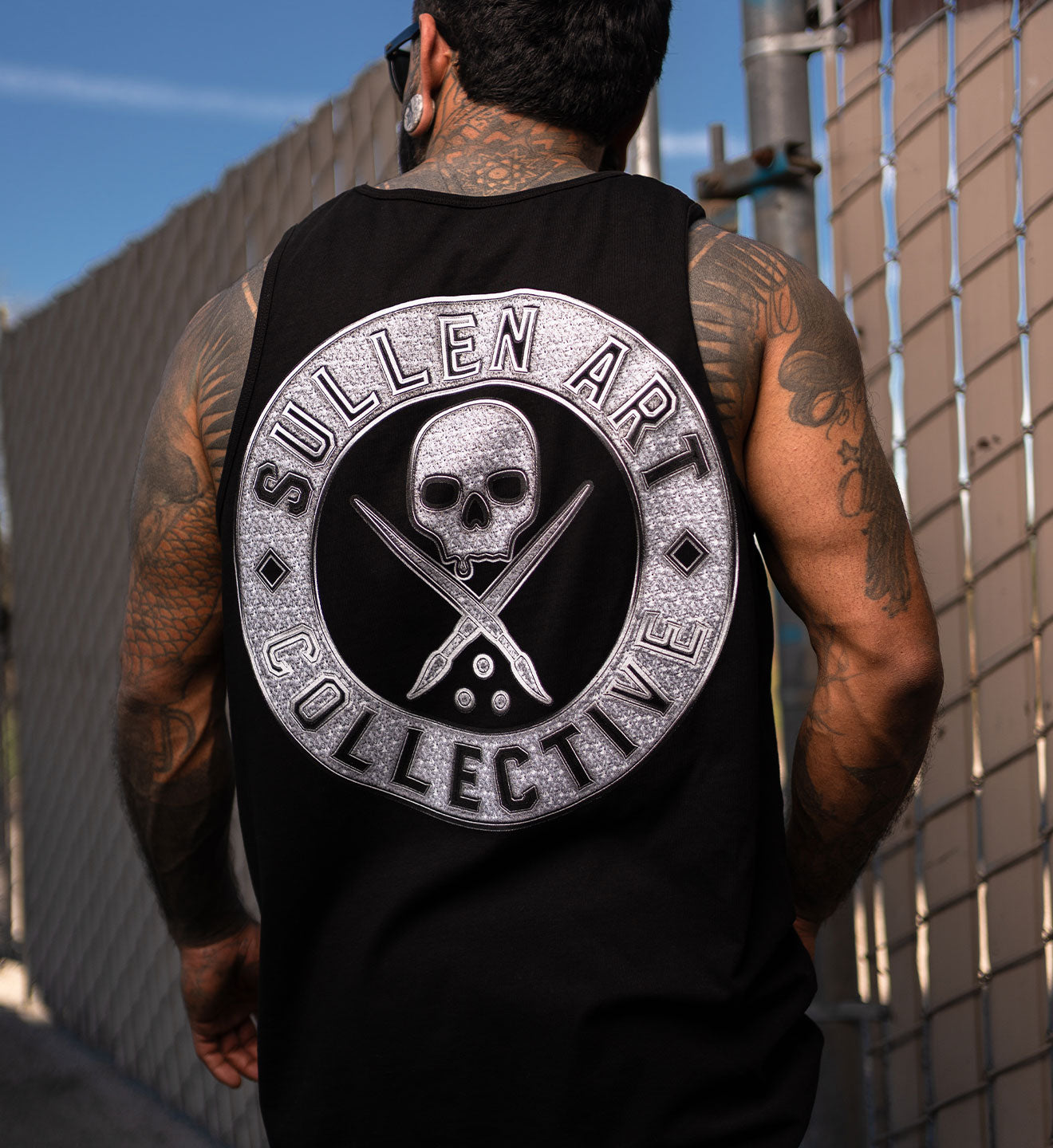 Person wearing a black tank top with 'Sullen Art Collective' logo on the back, standing against a textured wall.
