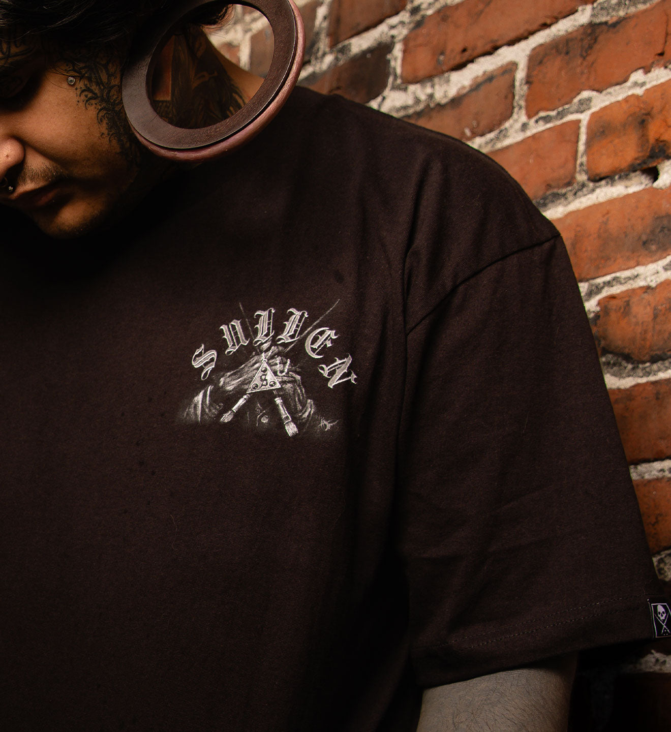 Person wearing a black t-shirt with a logo against a brick wall.