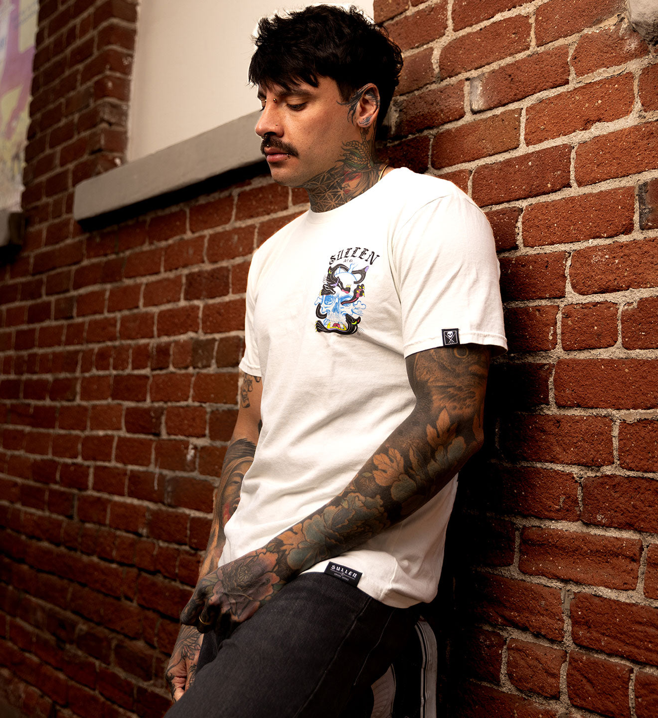 Man with tattoos leaning against a brick wall wearing a white t-shirt.