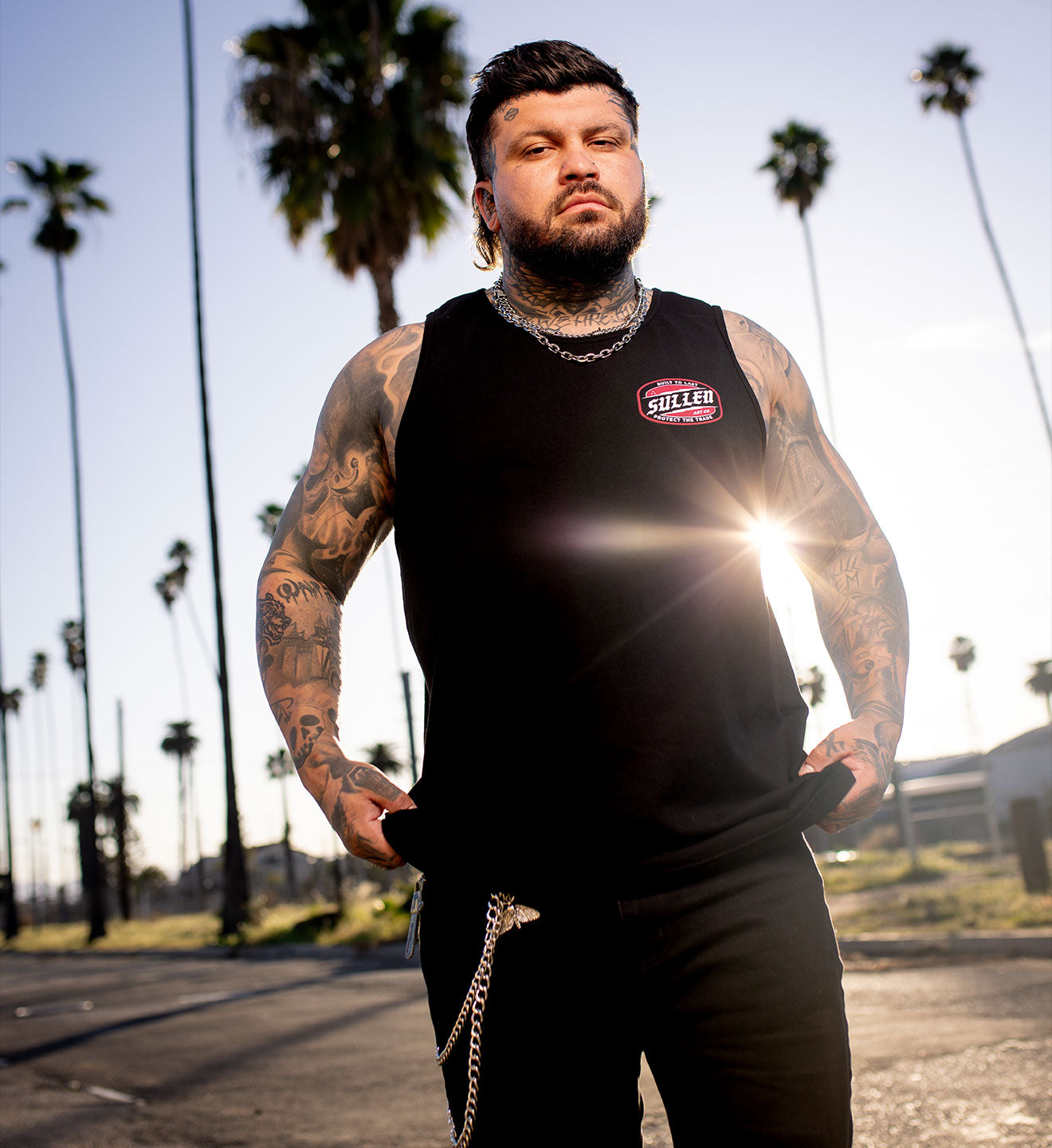 Man with tattoos wearing a black tank top with a logo, standing in a sunlit street with palm trees.