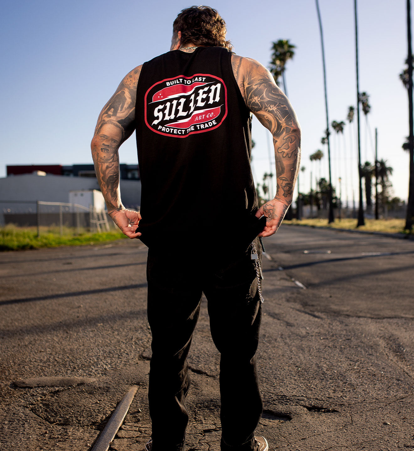 Man wearing a black sleeveless shirt with a logo, standing on a road with palm trees in the background.