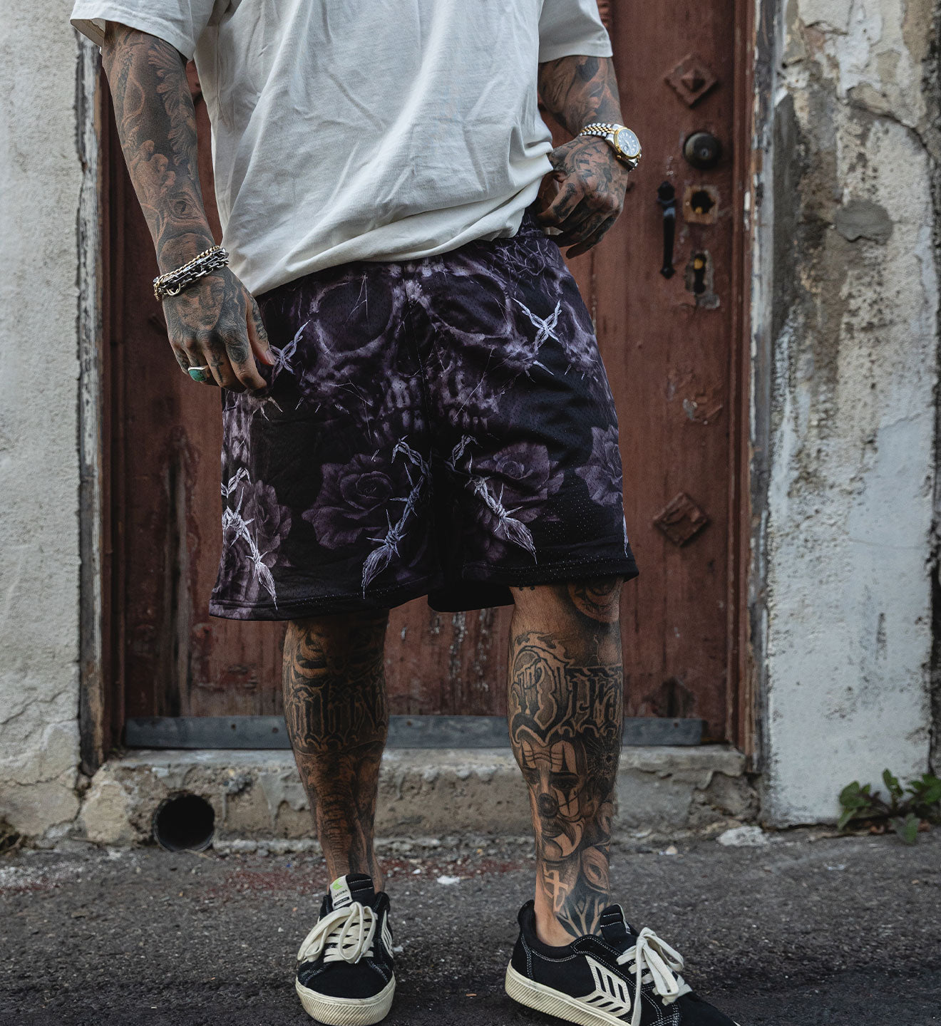 Person wearing a white t-shirt, purple patterned shorts, and black sneakers with tattoos on arms and legs, standing in front of a rustic door.