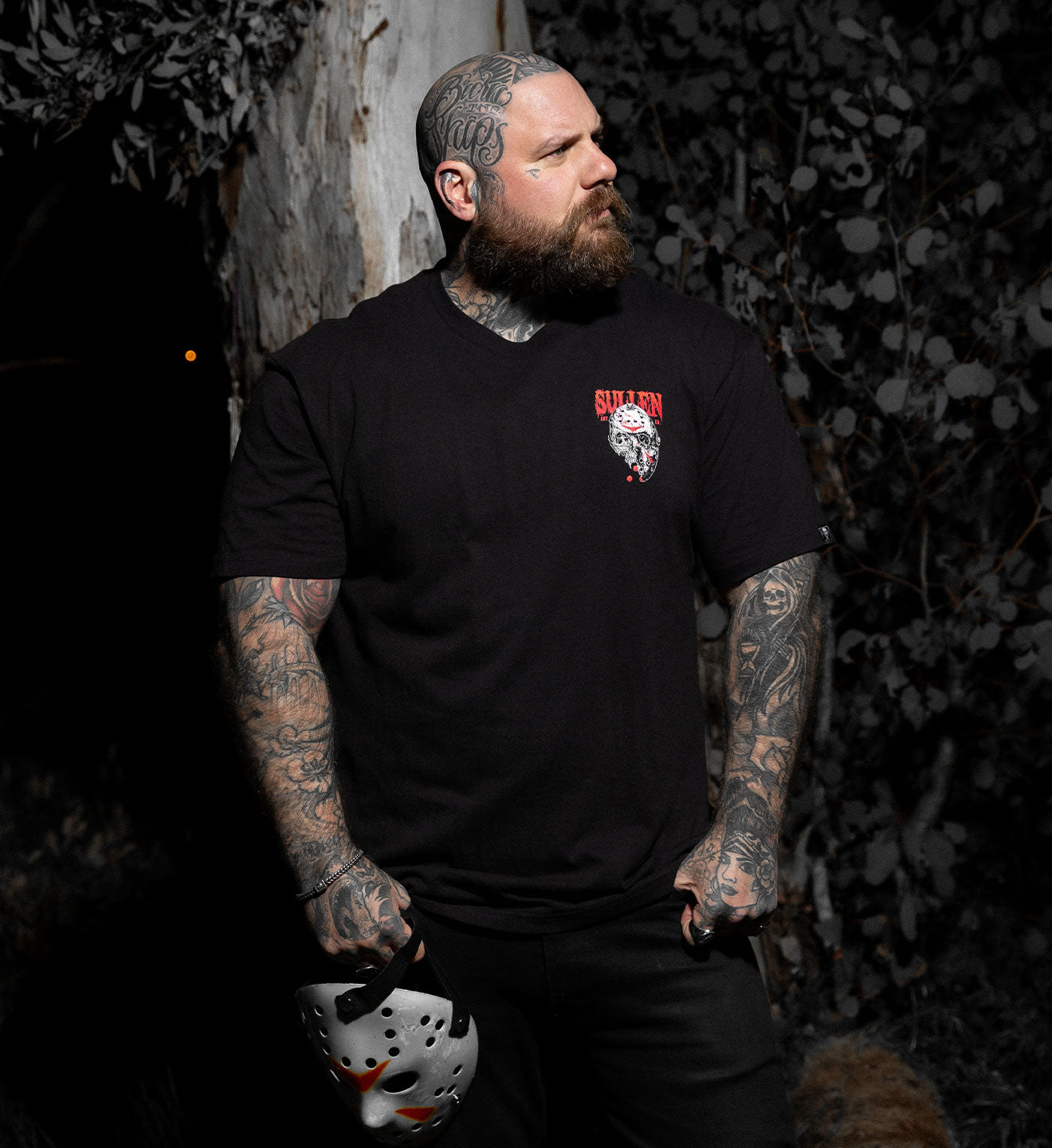 Man wearing a black t-shirt with a logo, holding a hockey mask, standing against a dark background.